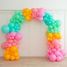 a rainbow colored balloon arch in front of a white wall with wood flooring and wooden floors