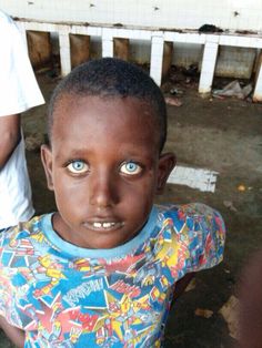 a young boy with blue eyes standing in front of a white fence and looking at the camera