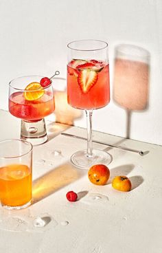 two glasses filled with drinks sitting next to oranges and strawberries on a table