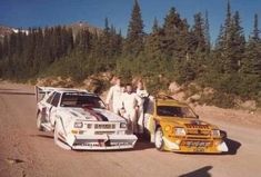 three men standing next to two cars on a dirt road