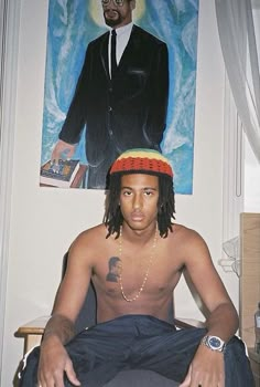 a man with dreadlocks sitting on a bed in front of a painting and wearing a hat