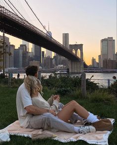 a man and woman sitting on a blanket in front of the brooklyn bridge at sunset