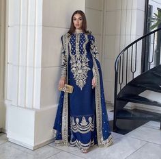 a woman standing in front of a staircase wearing a blue dress with gold embroidery on it