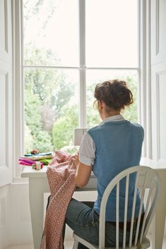 a woman sitting at a window sill working on her sewing project