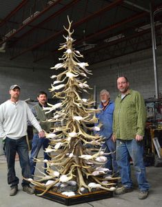 three men standing next to a large wooden christmas tree with antlers on it in a warehouse