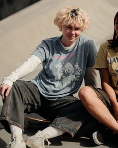 two young people sitting on the ground with one holding his skateboard and the other looking at the camera