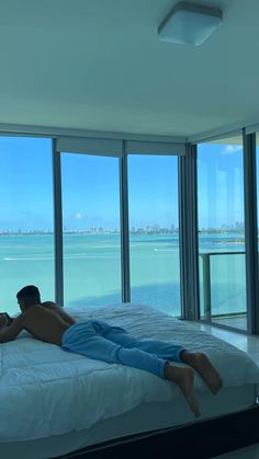 a man laying on top of a bed in front of large windows overlooking the ocean