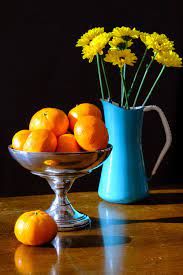 oranges and daisies in a glass bowl on a table next to a blue pitcher