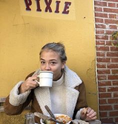 a woman sitting at a table drinking from a coffee cup while holding a plate with food in front of her