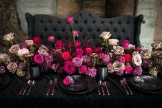 the table is set with black plates and silverware, pink flowers in vases