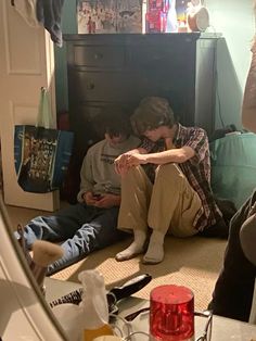 three young men sitting on the floor looking at their cell phones in front of a mirror