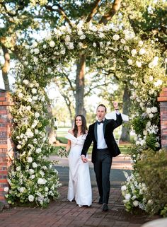 a bride and groom walking through an archway with white flowers on the arbor behind them