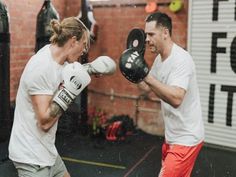 two men in white shirts and red shorts boxing with gloves on their hands, while one man looks at the camera