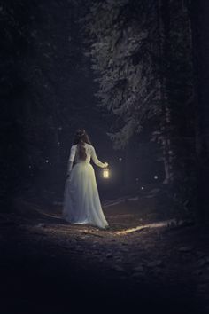 a woman in a white dress is walking through the woods at night holding a lantern