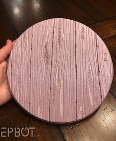 a hand holding a pink wooden coaster on top of a wood floor