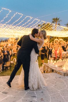 a bride and groom kissing in front of an outdoor wedding reception with string lights on the ceiling