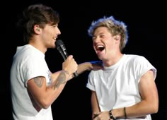 two men are laughing while one holds a microphone and the other is singing into it