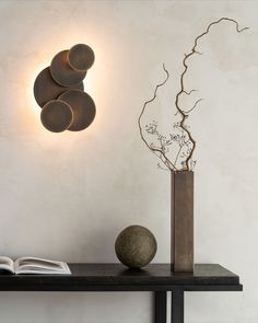 a vase with some branches in it next to a wall light and a book on a table