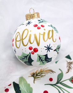 a christmas ornament with holly and gold lettering on it, sitting on top of wrapping paper