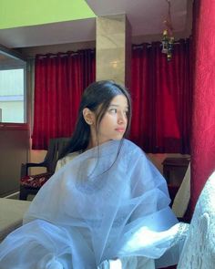 a woman in a white dress sitting on a couch with red curtains behind her and looking off into the distance