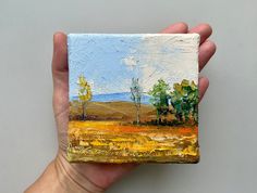 a person holding up a small painting in their hand with trees on the other side