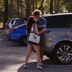 two people standing next to each other in front of a parked car and one person is kissing the woman's head