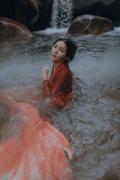 a woman sitting in the water with her hand up to her face and wearing an orange dress