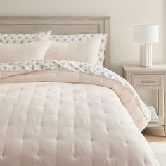 a bed with pink comforter and pillows in a white room next to a night stand