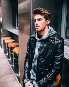 a man in a leather jacket leaning against a wall next to some chairs and tables