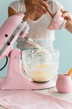 a woman pouring batter into a bowl on top of a table next to a pink mixer