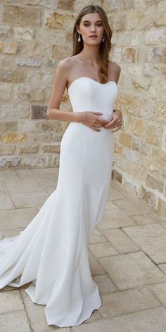 a woman standing in front of a stone wall wearing a white dress and holding her hand on her hip