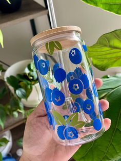 a hand holding a glass with blue flowers and leaves on it, in front of some plants