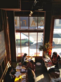 people are sitting at tables in a coffee shop looking out the window and writing on paper