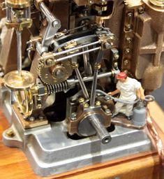 a clock made out of mechanical parts on a wooden table with a toy man standing next to it