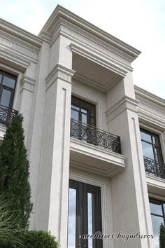 a tall white building with black doors and balcony railings on the second floor, next to a tree