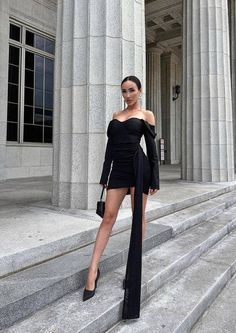 a woman standing on steps in front of a building wearing a black off the shoulder dress