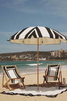 A picturesque beach setup with black-and-white striped umbrellas and matching chairs on a sandy shore. The ocean waves and distant coastal town create an inviting scene for seaside relaxation. Function Beauty, Pillow Lounger, Commercial Umbrellas, Coco Republic, Pool Lounger, Umbrella Shop, Beach Cabana, Beach Tent, Beach Shop