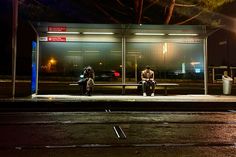 two people sitting on a bench in front of a bus stop at night with the lights on