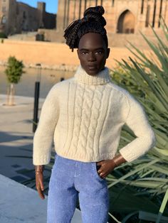 a doll is standing in front of a building with a large plant behind it and wearing a white sweater and blue pants