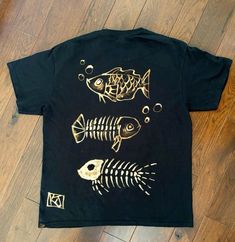 a black t - shirt with gold fish and skeleton bones on the front, sitting on a wooden floor