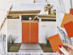 a person holding an orange card in front of a white house with red doors and paint swatches