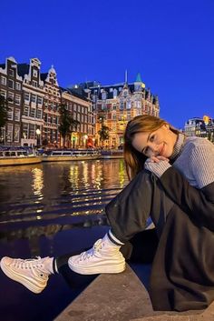 a woman sitting on the edge of a body of water in front of some buildings