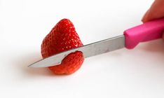 a strawberry being cut by a knife on a white surface with pink handle and two small strawberries in the foreground