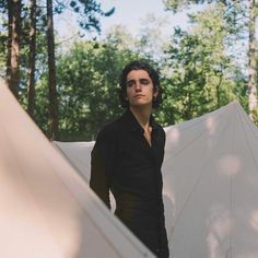 a man standing next to a white tent in the middle of a forest with trees behind him