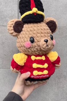 a hand holding a small teddy bear wearing a red and yellow shirt with a black hat on it's head