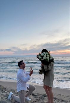 a man kneeling down next to a woman on top of a sandy beach near the ocean