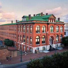 an old red brick building with green roof and people walking around the courtyard at sunset