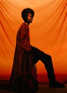 a man sitting on top of a chair in front of an orange curtain with his legs crossed