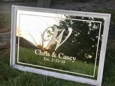 a glass sign sitting in the grass near a tree and building with a v on it's side