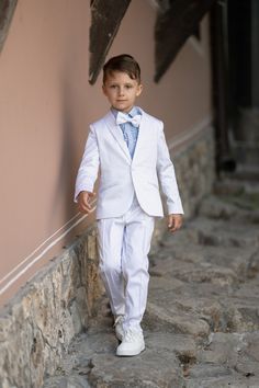 a young boy dressed in a white suit and bow tie walking down a stone walkway